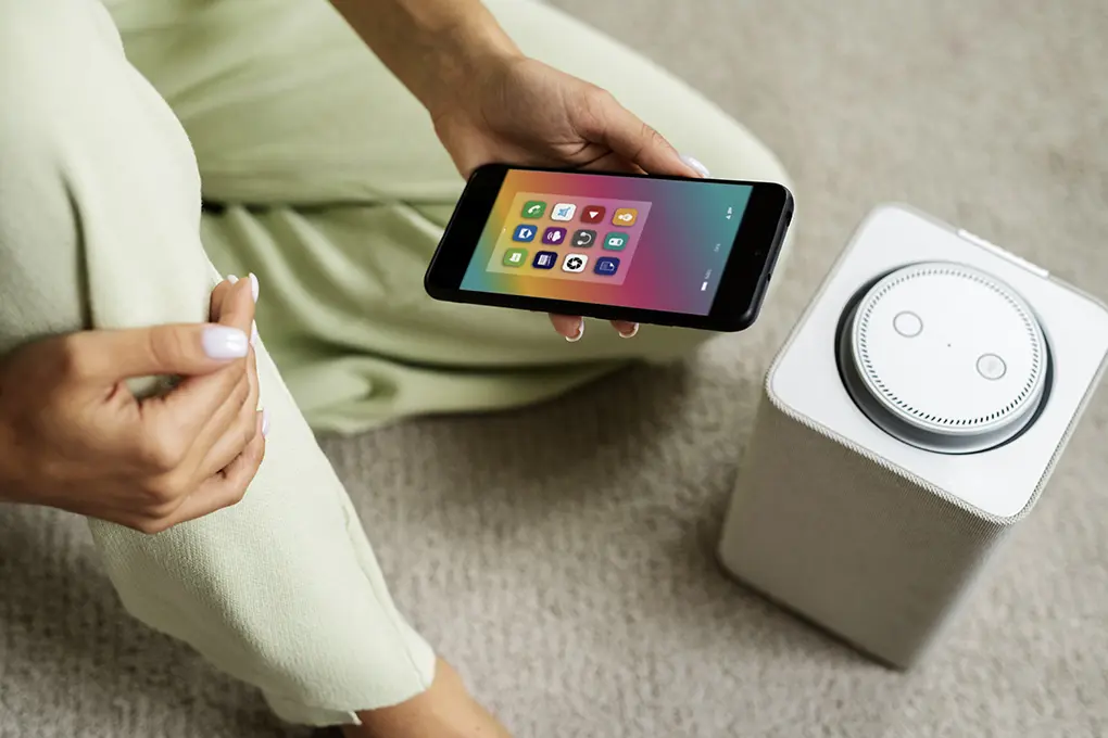 Person holding a smartphone displaying app icons, next to a smart speaker on a light-colored carpet