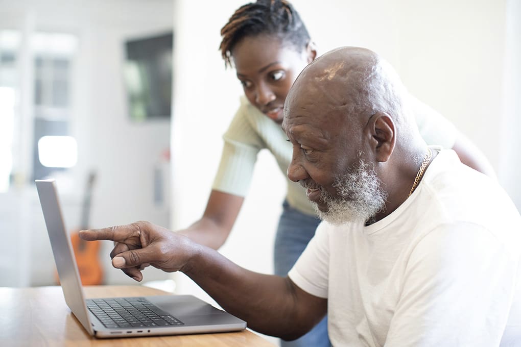 An elderly man in a white shirt points at a laptop screen as a woman leans over, smiling and assisting with cybersecurity tasks