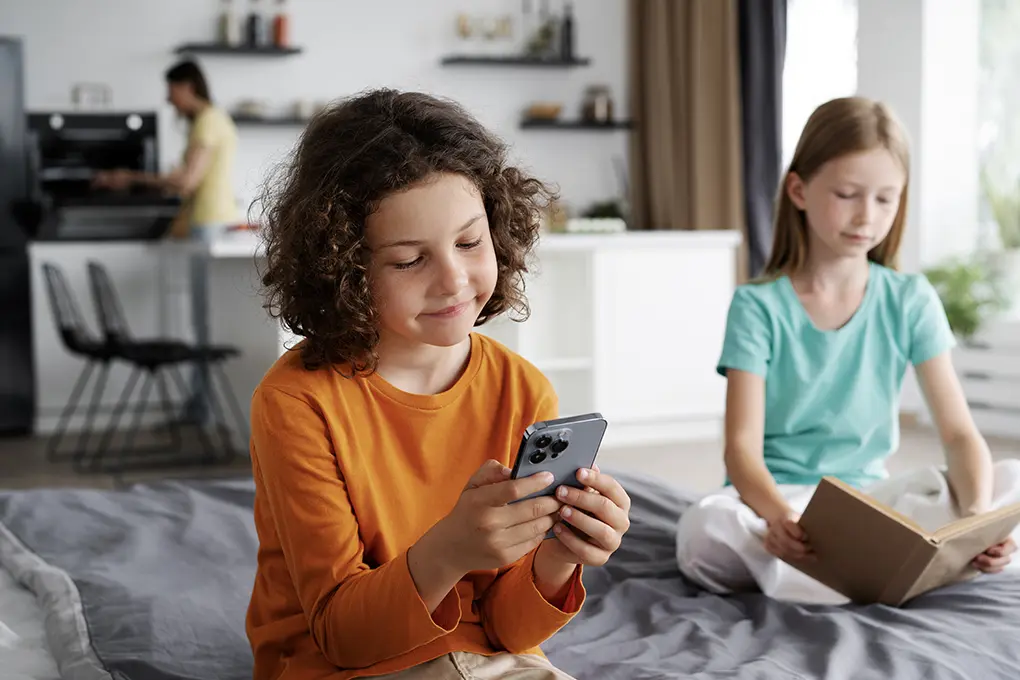 A boy in an orange shirt smiles while using a smartphone on a bed. Beside him, a girl in a teal shirt reads a book. A person is in the background, cooking