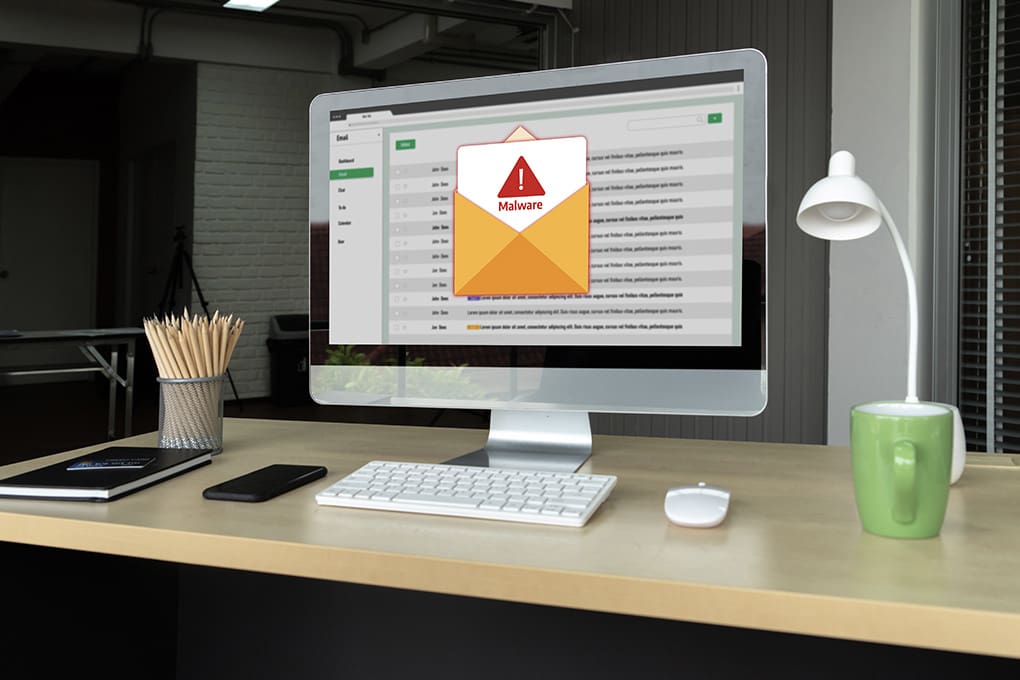 A desktop displays an email inbox on screen with a phishing email warning, set in a modern office with a keyboard, mouse, green mug, lamp, and pencil holder