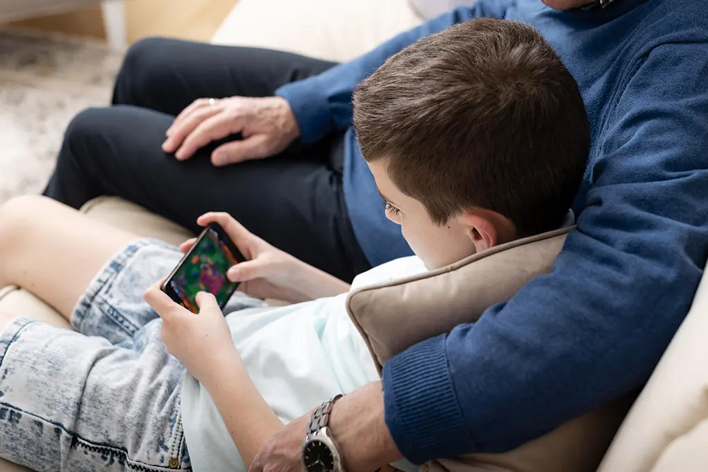 A boy and an adult relax on a couch & the boy, focused, playing a game on a smartphone