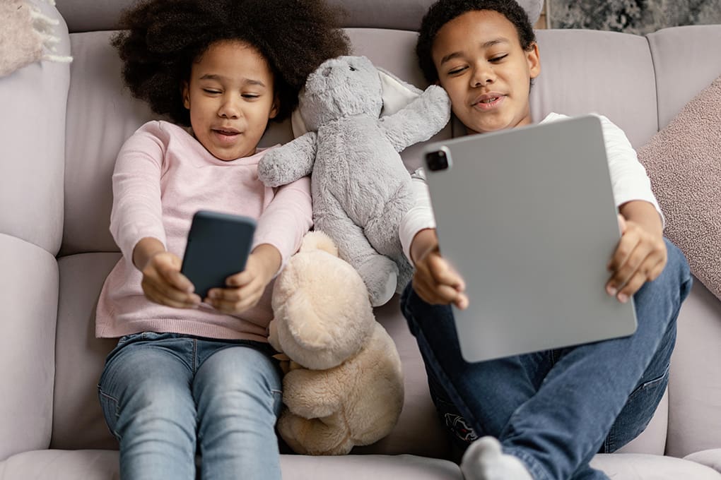 Two children are relaxing on a sofa, one holding a smartphone and the other an iPad